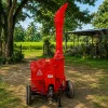 Red portable wood chipper on wheels with a tall curved chute, set in a rural yard with trees and fields in the background.