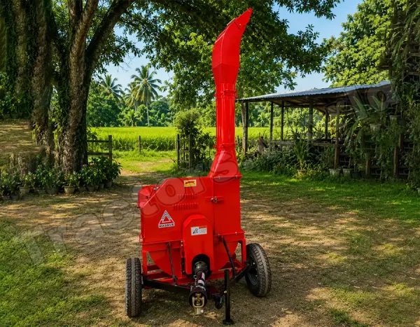 Red portable wood chipper on wheels with a tall curved chute, set in a rural yard with trees and fields in the background.