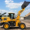 Yellow telehandler loader at a quarry raises its front bucket to move gravel with piles and conveyors in the background